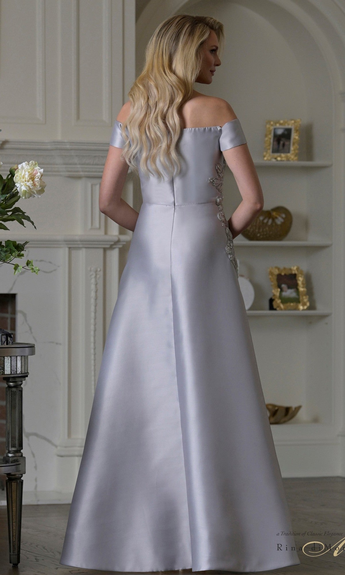 A woman with long blonde hair stands facing away in an elegant room, wearing the Rina Di Montella RD3501—an off-the-shoulder, floor-length silver Mikado formal dress with an a-line skirt.